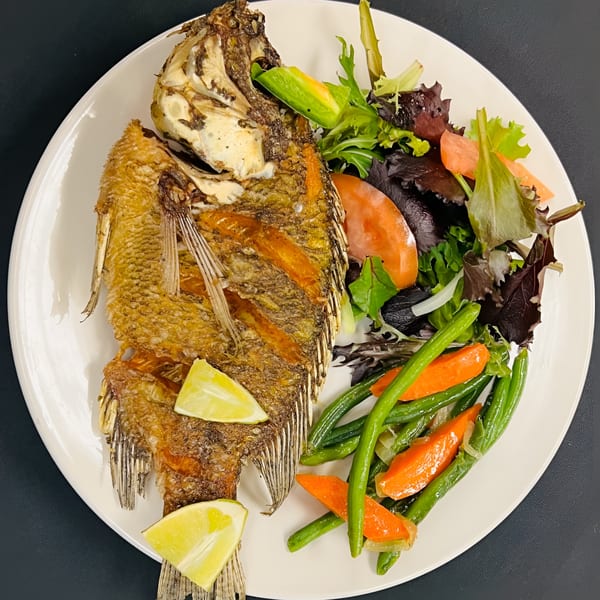 Whole fried fish with lime wedges, green beans, carrots, and a side salad on a white plate.