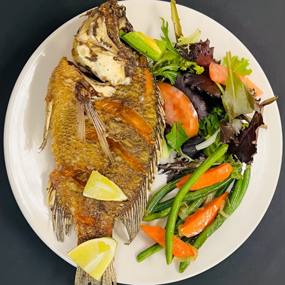 Whole fried fish with lime wedges, green beans, carrots, and a side salad on a white plate.